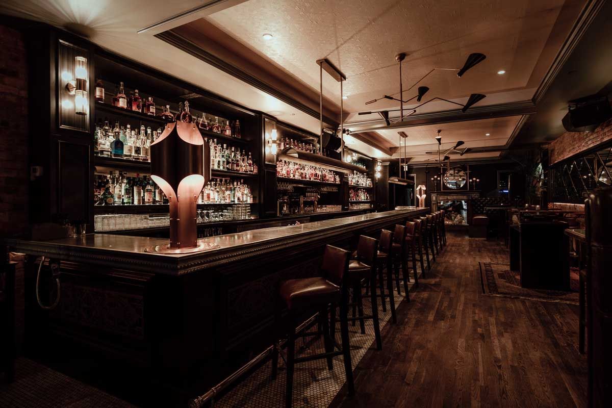 Dimly lit speakeasy-style cocktail bar interior with a long polished wood bar, leather stools, backlit shelves of spirits and warm pendant lighting, cozy urban nightlife vibe