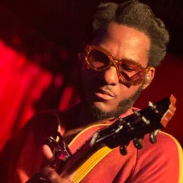 Close-up of a guitarist wearing vintage amber sunglasses and a red shirt, focused while playing an electric guitar on a dimly lit live music stage with warm red lighting.