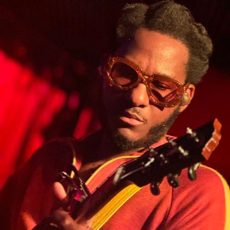 Close-up of a guitarist wearing vintage amber sunglasses and a red shirt, focused while playing an electric guitar on a dimly lit live music stage with warm red lighting.