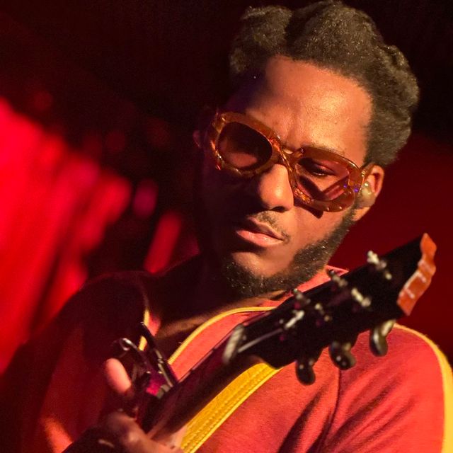 Close-up of a guitarist wearing vintage amber sunglasses and a red shirt, focused while playing an electric guitar on a dimly lit live music stage with warm red lighting.