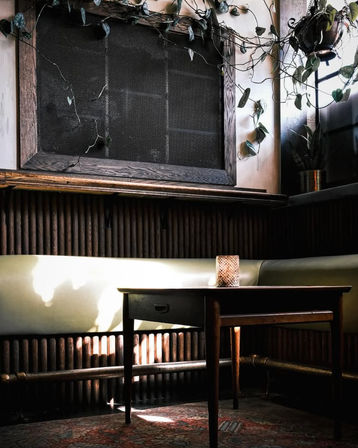 Cozy urban cafe corner with a sunlit wooden table and small drawer, pale green booth seating, hanging leafy vines and a potted plant, framed mesh wall, ribbed wood paneling and exposed radiator pipes over a patterned rug.