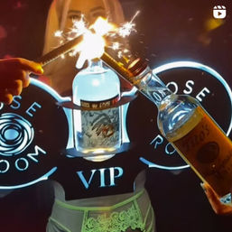 VIP nightclub bottle service: sparklers bursting over a lit liquor bottle held between hands in a neon-logo booth, celebratory party vibe
