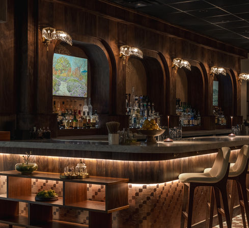 Dimly lit upscale cocktail bar interior with curved wood arches, marble countertop, crystal wall sconces, backlit bottles, and cushioned bar stools
