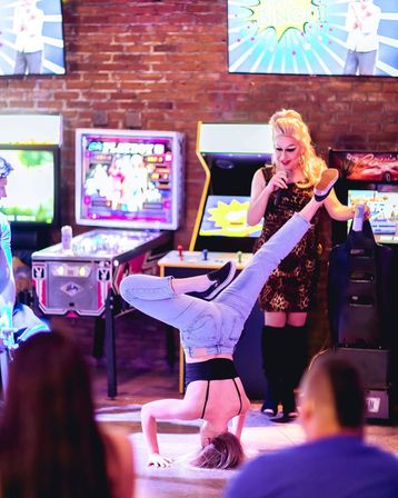 Person doing a headstand in jeans on an arcade floor while a blonde stage performer with a microphone entertains beside pinball and retro arcade machines at a lively arcade bar nightlife event