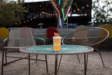 Peach-colored cocktail with dried citrus garnish on a woven round patio table, surrounded by colorful metal lounge chairs and soft string lights in a blurred outdoor bar setting
