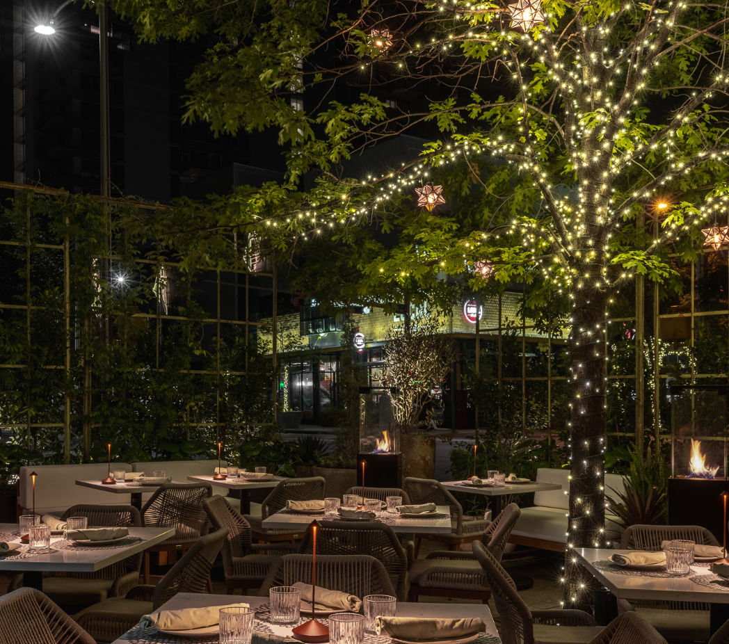 Cozy urban outdoor dining patio at night with a tree wrapped in twinkling string lights and candlelit tables set for dinner.