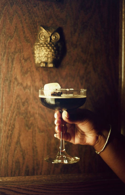 Hand holding a coupe glass with a dark coffee-colored cocktail and a large ice cube, set against a moody wood-paneled wall with a brass owl wall ornament.