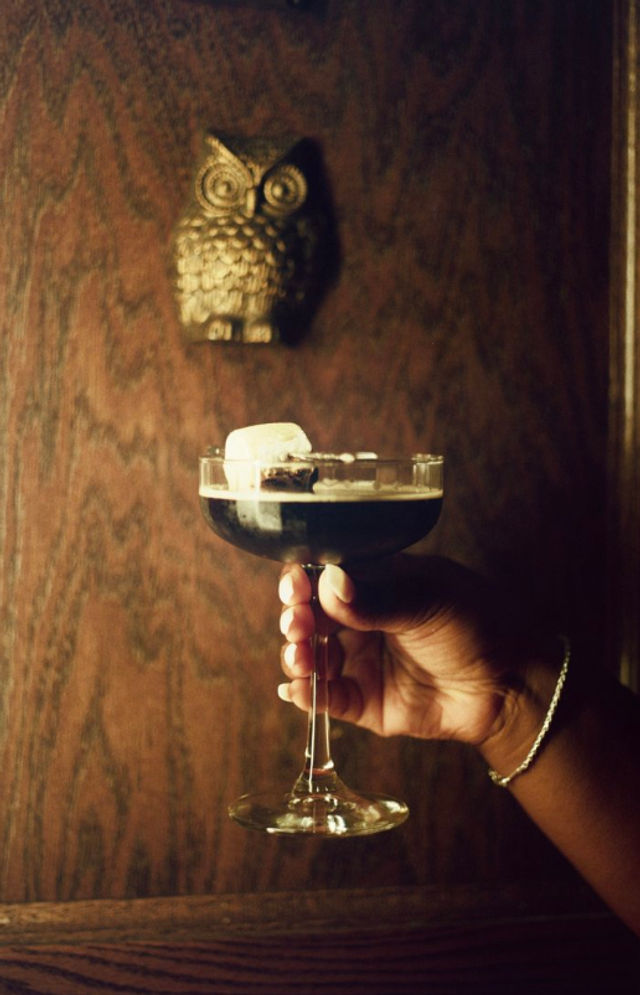 Hand holding a coupe glass with a dark coffee-colored cocktail and a large ice cube, set against a moody wood-paneled wall with a brass owl wall ornament.