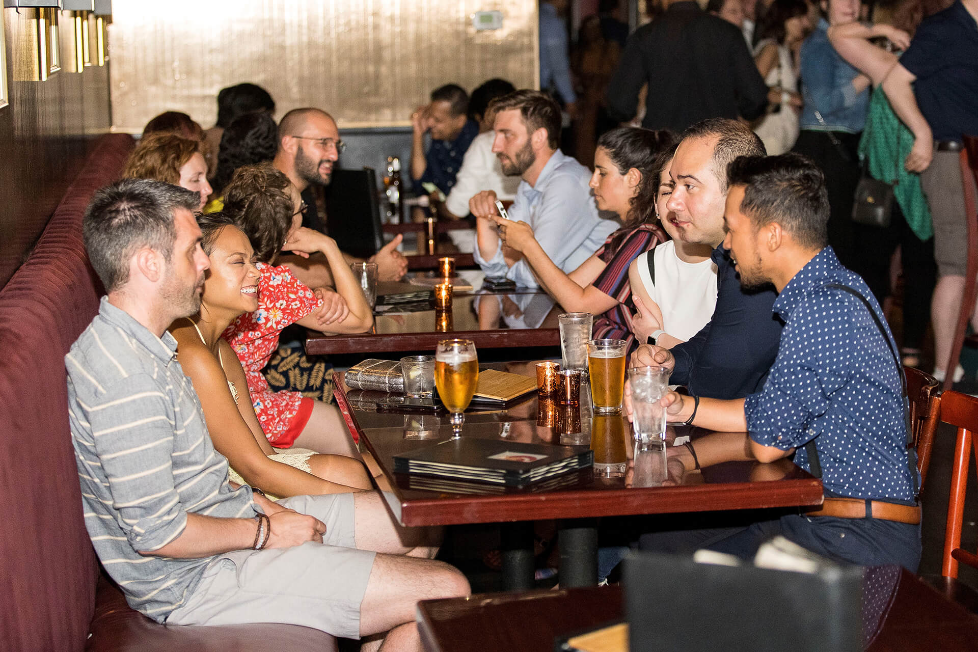 Lively urban bar interior with groups seated along a banquette, friends laughing and drinking beers and cocktails at wooden tables during a busy evening nightlife scene