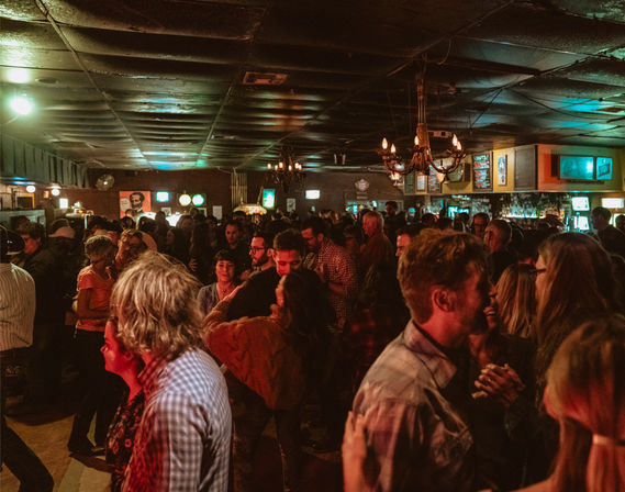 Crowded dimly lit bar interior with people dancing and chatting on a lively night, warm red and teal lighting, chandeliers overhead and a busy bar in the background.
