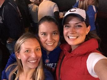 Three smiling women taking a selfie at a crowded sports bar, two wearing Chicago Cubs gear for a fun night out