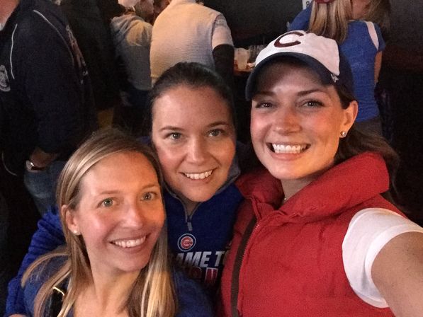 Three smiling women taking a selfie at a crowded sports bar, two wearing Chicago Cubs gear for a fun night out