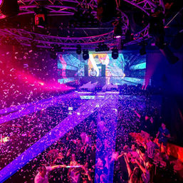 Packed nightclub dance floor with confetti, purple-blue laser beams and LED stage screens behind a DJ booth — energetic electronic music club scene.