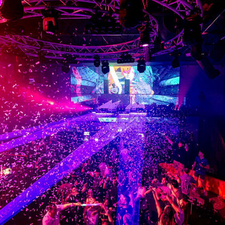 Packed nightclub dance floor with confetti, purple-blue laser beams and LED stage screens behind a DJ booth — energetic electronic music club scene.