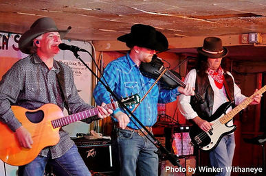 Three-piece country band in cowboy hats performing live with acoustic guitar, fiddler and electric bass on a small wooden venue stage