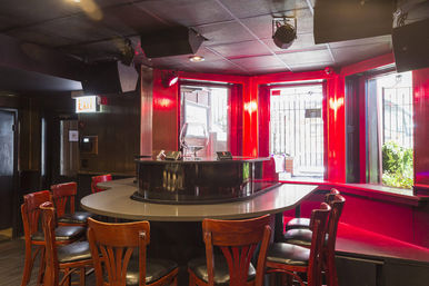 Cozy retro bar interior with glossy red walls and bay windows, a curved gray counter with a central raised service island, wooden bar stools, and stage lights overhead — intimate urban lounge vibe.