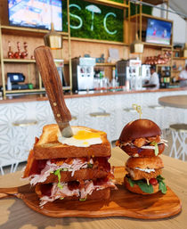 Knife-stabbed club sandwich with bacon, turkey, lettuce and a sunny-side-up egg on toasted bread, served on a wooden board next to two mini sliders at a bright brunch bar.