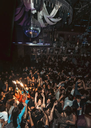 Packed nightclub dance floor with cheering crowd under a disco ball, bottle service sparklers lighting a lively late-night party scene