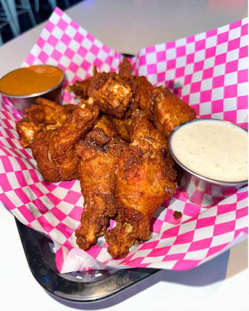 Crispy golden fried chicken wings in a pink checkered paper-lined basket with creamy ranch and orange dipping sauce — casual American pub-style appetizer.