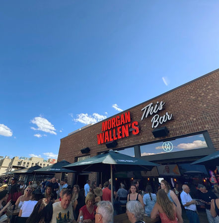 Bustling outdoor bar patio on a sunny day, crowds gathered under umbrellas in front of a brick facade with red neon and script signage.