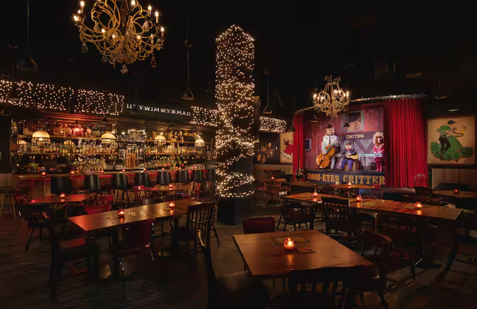 Cozy dimly lit bar and live-music venue with a small stage framed by red curtains, chandeliers and string lights wrapped around a central pillar, wooden tables with candlelit votives, and a stocked back bar with stools.