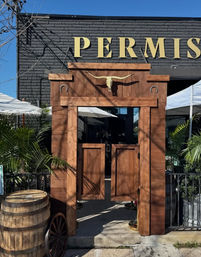 Western-style wooden saloon entrance with swinging batwing doors, longhorn skull and horseshoe accents, barrel and wagon wheel by an outdoor patio under a bright blue sky.