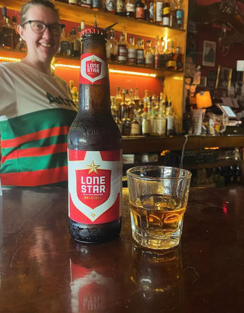 Chilled Lone Star beer bottle and small whiskey glass on a polished bar counter, blurred backbar bottles and a smiling bartender in a cozy pub setting.