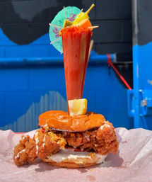 Crispy spicy fried chicken sandwich with mayo on a toasted bun, pierced by a tall chili-rimmed michelada garnished with fruit and a paper umbrella, photographed against a bright blue urban mural.