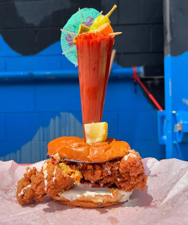 Crispy spicy fried chicken sandwich with mayo on a toasted bun, pierced by a tall chili-rimmed michelada garnished with fruit and a paper umbrella, photographed against a bright blue urban mural.