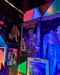 Neon-lit venue wall covered in colorful retro posters and artwork: framed 'Adventures in Babysitting' movie poster, chalk-style horse drawing, blue-tinted sequined performer portrait, vintage vinyl and a playful dog poster, all glowing purple, teal and magenta.