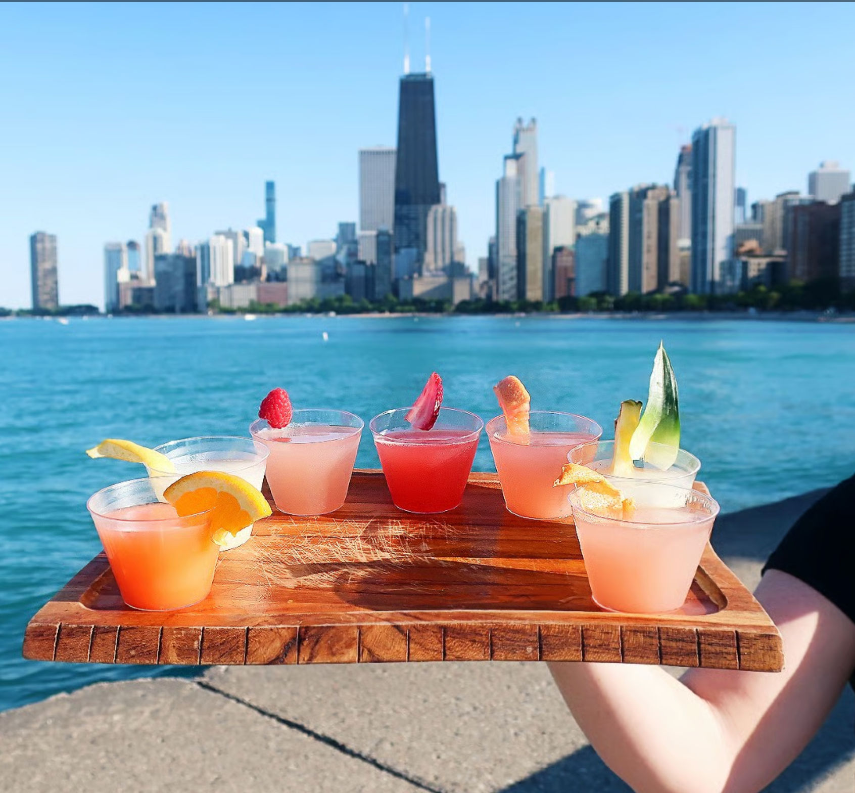Wooden flight of colorful mini cocktails garnished with citrus and berries held over Lake Michigan with the sunny Chicago skyline in the background