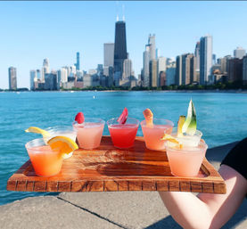 Wooden flight of colorful mini cocktails garnished with citrus and berries held over Lake Michigan with the sunny Chicago skyline in the background