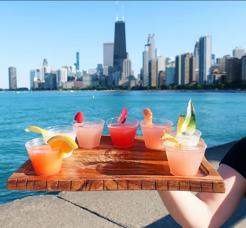 Wooden flight of colorful mini cocktails garnished with citrus and berries held over Lake Michigan with the sunny Chicago skyline in the background