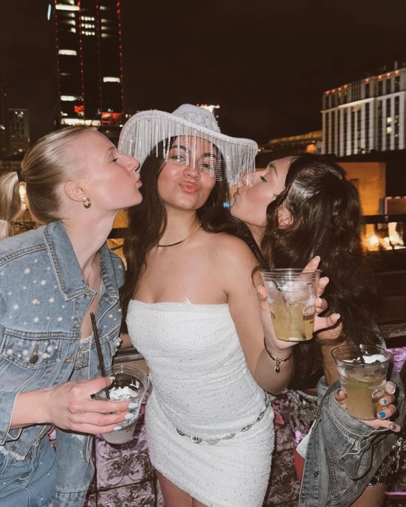 Three friends at a downtown rooftop party at night — woman in a white beaded cowgirl hat and strapless dress flanked by pals kissing her cheeks while holding cocktails with city skyline lights behind them.