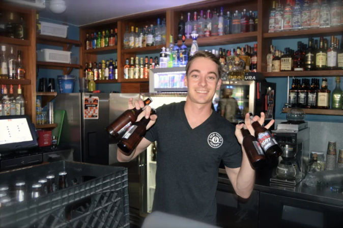 Smiling bartender cheerfully holding several beer bottles by the neck behind a busy pub counter, with liquor shelves, a refrigerated display, and bar equipment in the background.