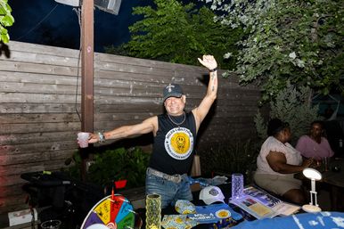 Smiling person in a cap and sleeveless shirt raises arms behind a vendor table with merch and a colorful prize wheel at an outdoor evening community patio event.