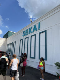 Sunny outdoor storefront with teal trim and oversized signage, people in colorful summer outfits waiting on the asphalt under a bright blue sky with clouds.