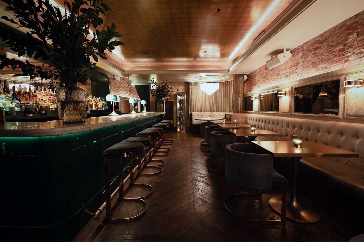 Cozy upscale cocktail bar interior with a marble-topped green bar, brass stools, tufted velvet banquettes, round wooden tables with candles, exposed brick walls and warm ambient lighting