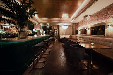 Cozy upscale cocktail bar interior with a marble-topped green bar, brass stools, tufted velvet banquettes, round wooden tables with candles, exposed brick walls and warm ambient lighting