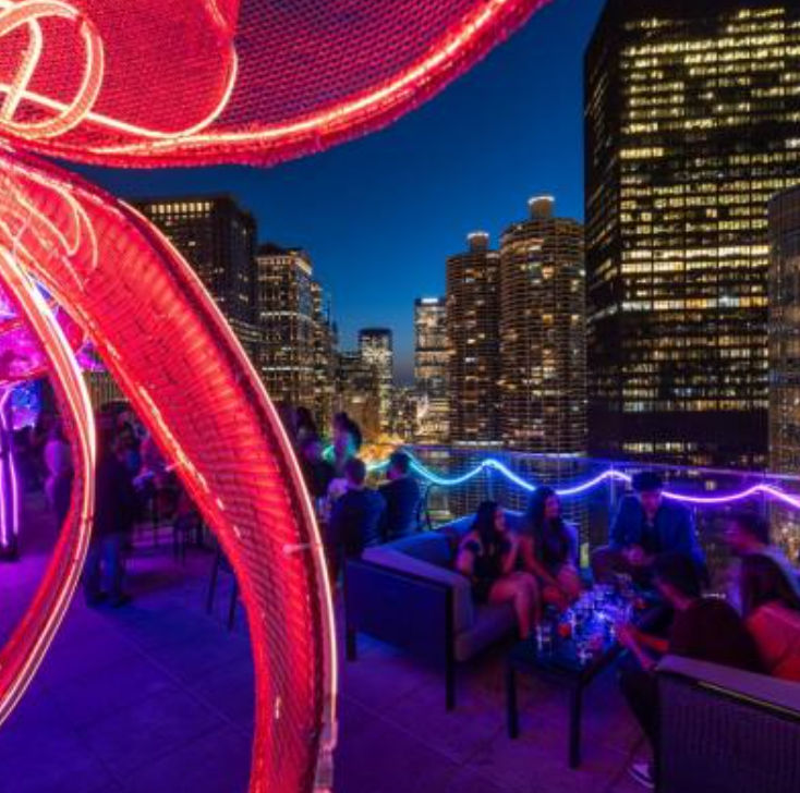 Vibrant rooftop lounge at night — glowing red neon sculpture in the foreground, people socializing on sofas under purple-blue string lights with a twinkling downtown skyscraper skyline beyond.