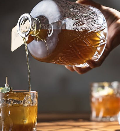 Hand pouring amber whiskey from a cut-glass decanter into an ice-filled lowball cocktail glass garnished with a herb pick on a wooden table, blurred second glass in the background