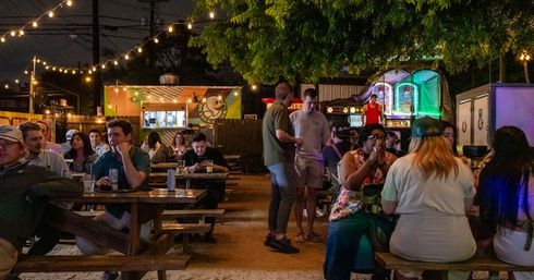 Nighttime outdoor patio bar scene with string lights, picnic tables of people eating and drinking, a colorful food truck and illuminated arcade games under trees, lively urban hangout