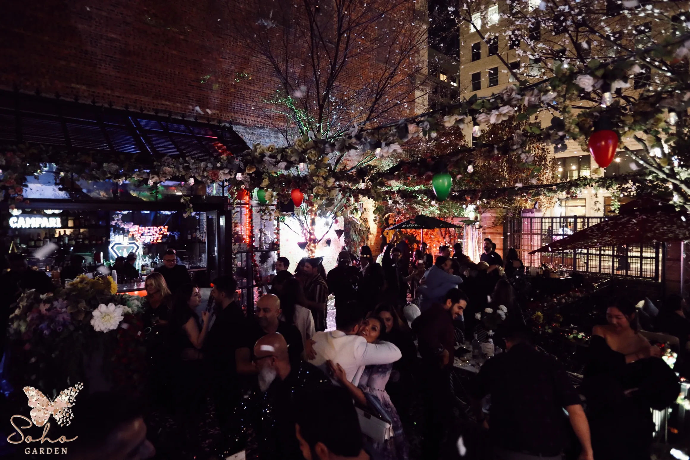 Festive urban rooftop garden patio at night with twinkling string lights, floral canopy and red and green balloons, crowds mingling near a bar against brick buildings.