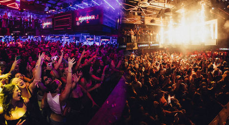 Packed two-level nightclub with a cheering dance crowd and balcony, colorful neon and LED lights, stage pyrotechnics and high-energy party atmosphere.