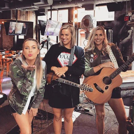 Three women acoustic musicians pose on a small urban bar stage — one blows a kiss, one holds a mandolin, and one holds an acoustic guitar ready for live music.