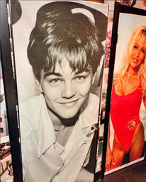 Black-and-white close-up poster of a smiling young man on a folding indoor display panel beside a colorful poster of a blonde woman in a red one-piece swimsuit — retro-style wall art.