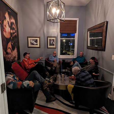 Cozy gray lounge with a geometric pendant light and large portrait, six people in armchairs chatting around a round coffee table while city lights glow outside the window.