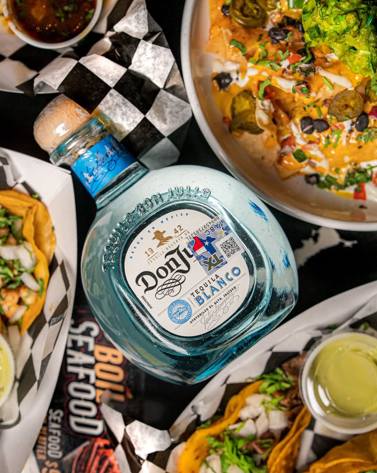 Overhead shot of a Mexican street-food spread featuring a blue glass bottle of blanco tequila surrounded by tacos, loaded nachos with guacamole, and dipping sauces on checkered paper