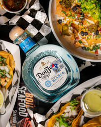 Overhead shot of a Mexican street-food spread featuring a blue glass bottle of blanco tequila surrounded by tacos, loaded nachos with guacamole, and dipping sauces on checkered paper