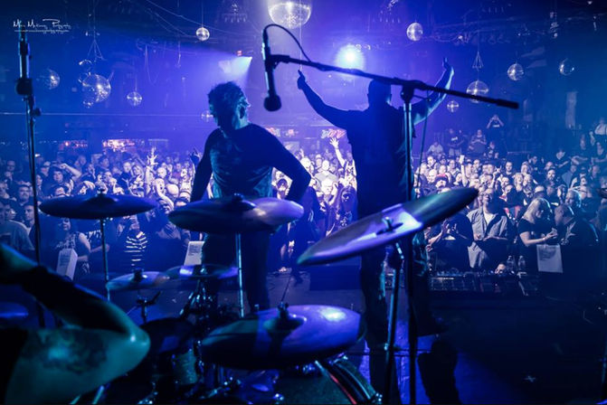 View from behind a drum kit at a packed indoor concert — silhouetted performers on stage, disco balls and blue-purple lighting, cheering crowd.
